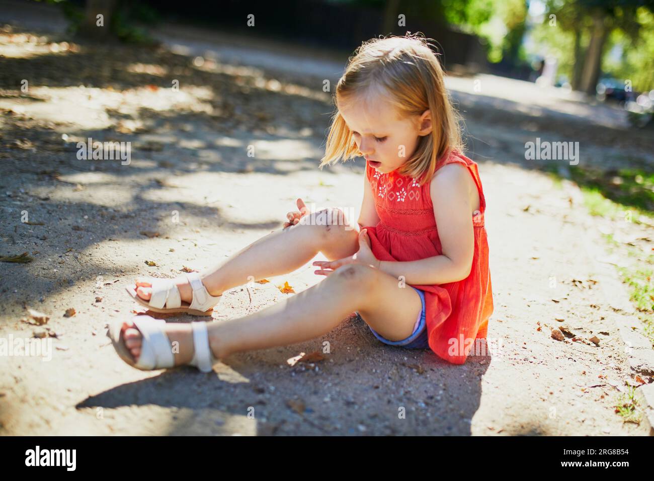 Cute little girl sitting on the ground after falling down. Child getting hurt while walking in park. Kid looking at her boo-boo Stock Photo