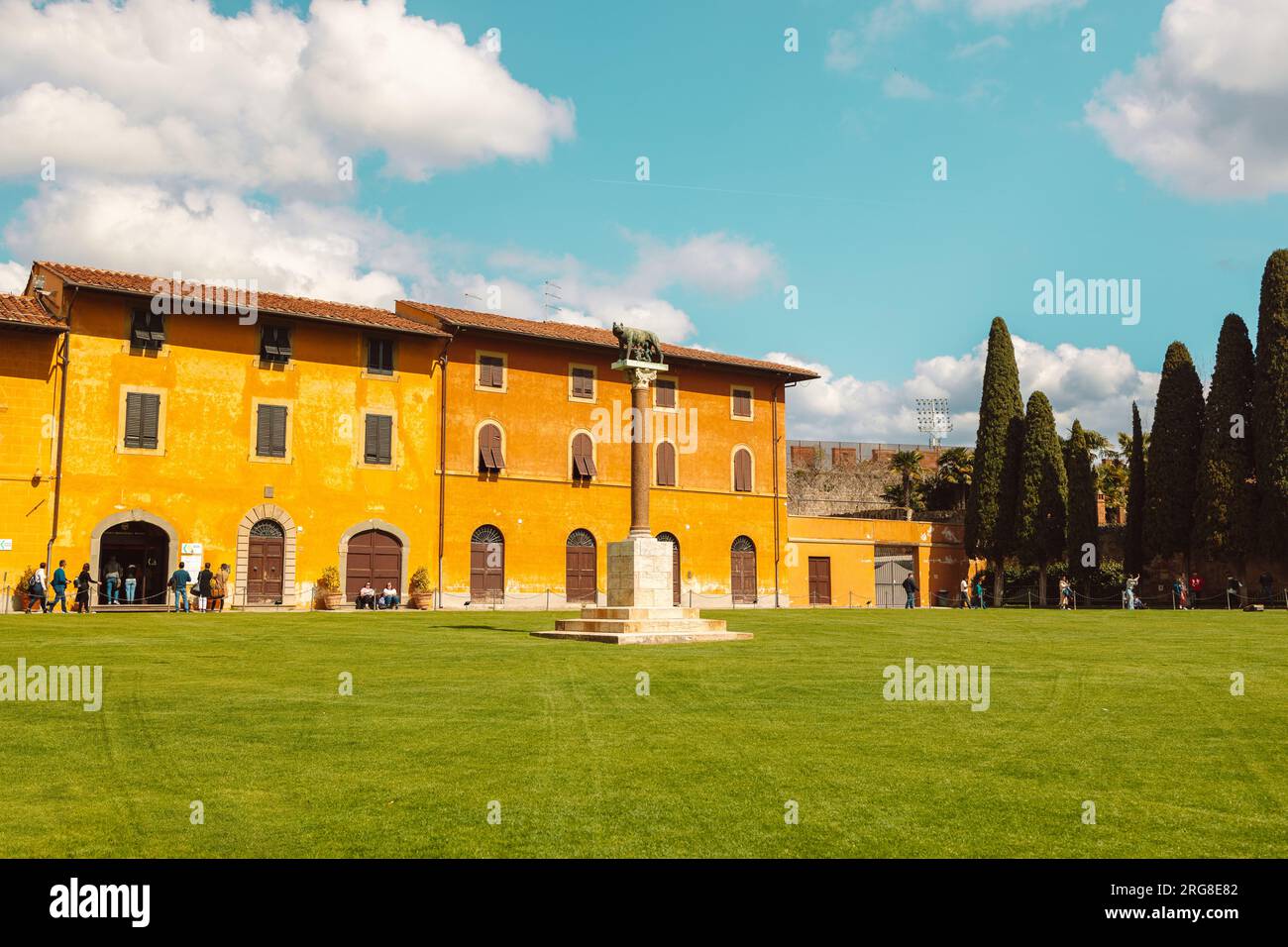 Pisa, Italy - March 18, 2023: Palazzo dell'Opera palace, Angelo Caduto statue, Lupa capitolina monument on square with green grass lawn, blue sky Stock Photo