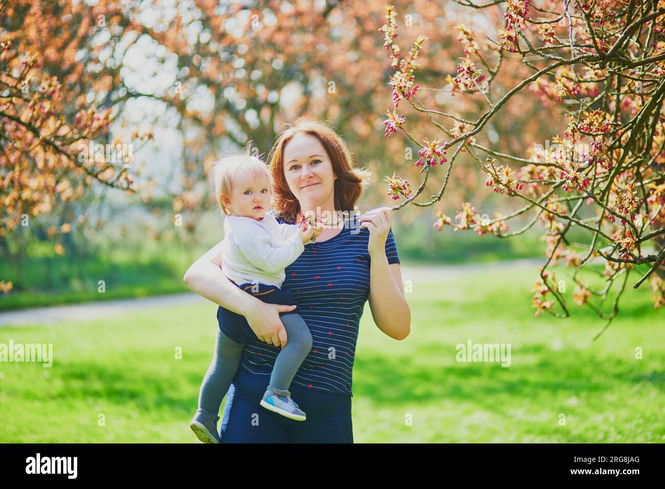 Young woman holding her 1 year old baby girl in park on a spring day. Mother and daughter enjoying cherry blossom season. Family activities with kids Stock Photo