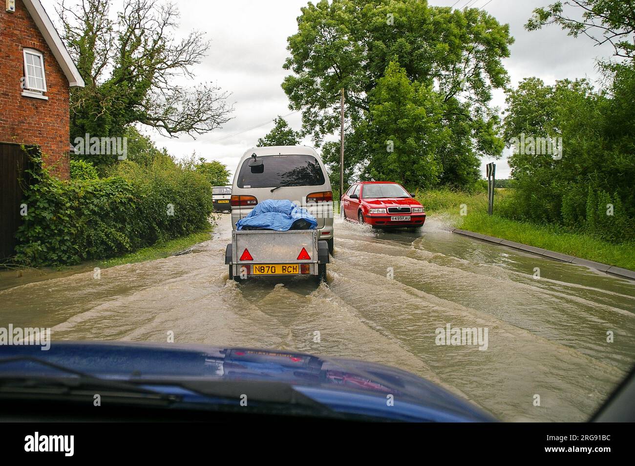 Driving through flooded road with cars. Driver's view from inside car driven through rainwater flood in Lincolnshire country road near property. July Stock Photo