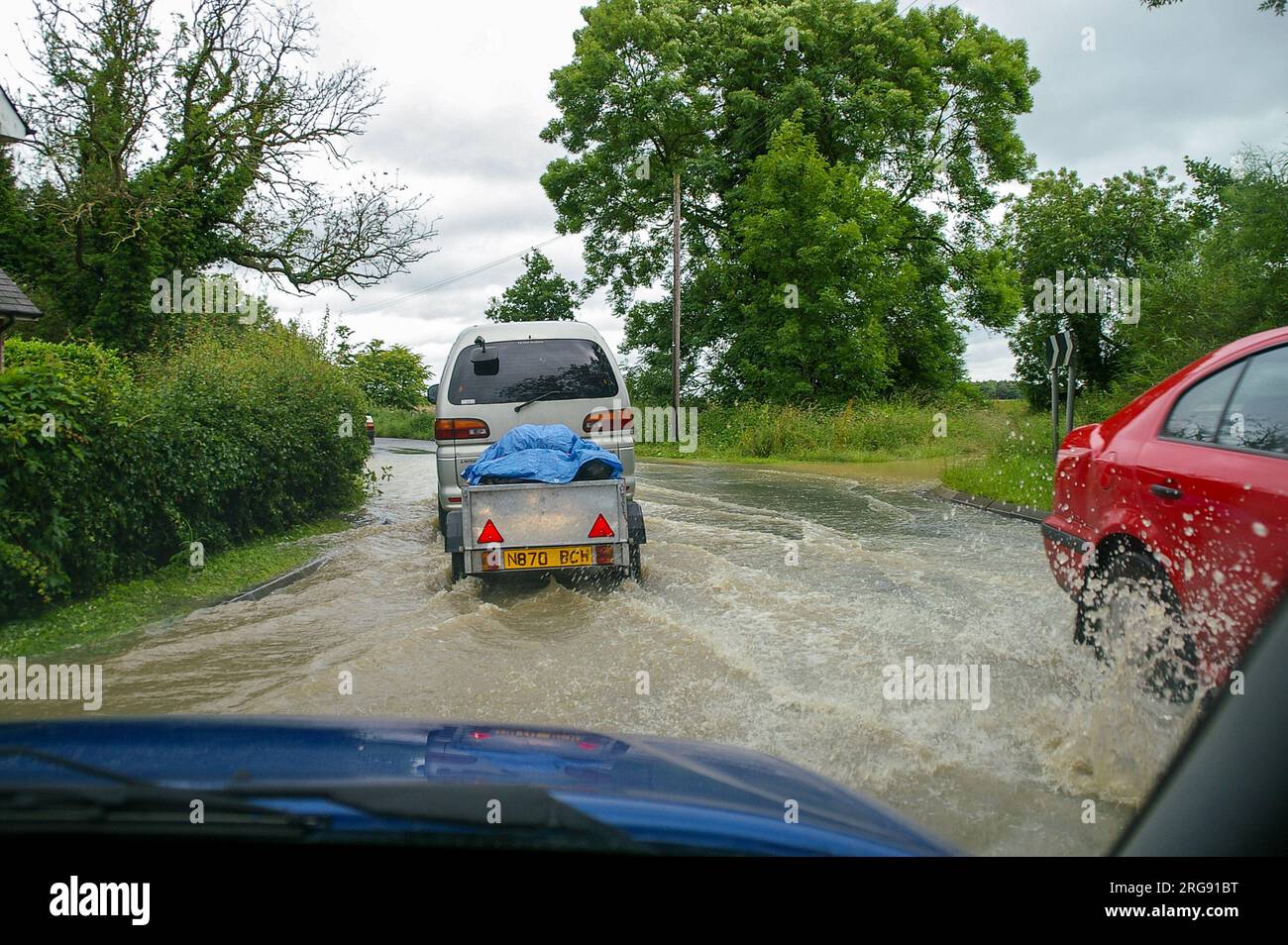 Driving through flooded road with cars. Driver's view from inside car driven through rainwater flood in Lincolnshire country road near property. July Stock Photo