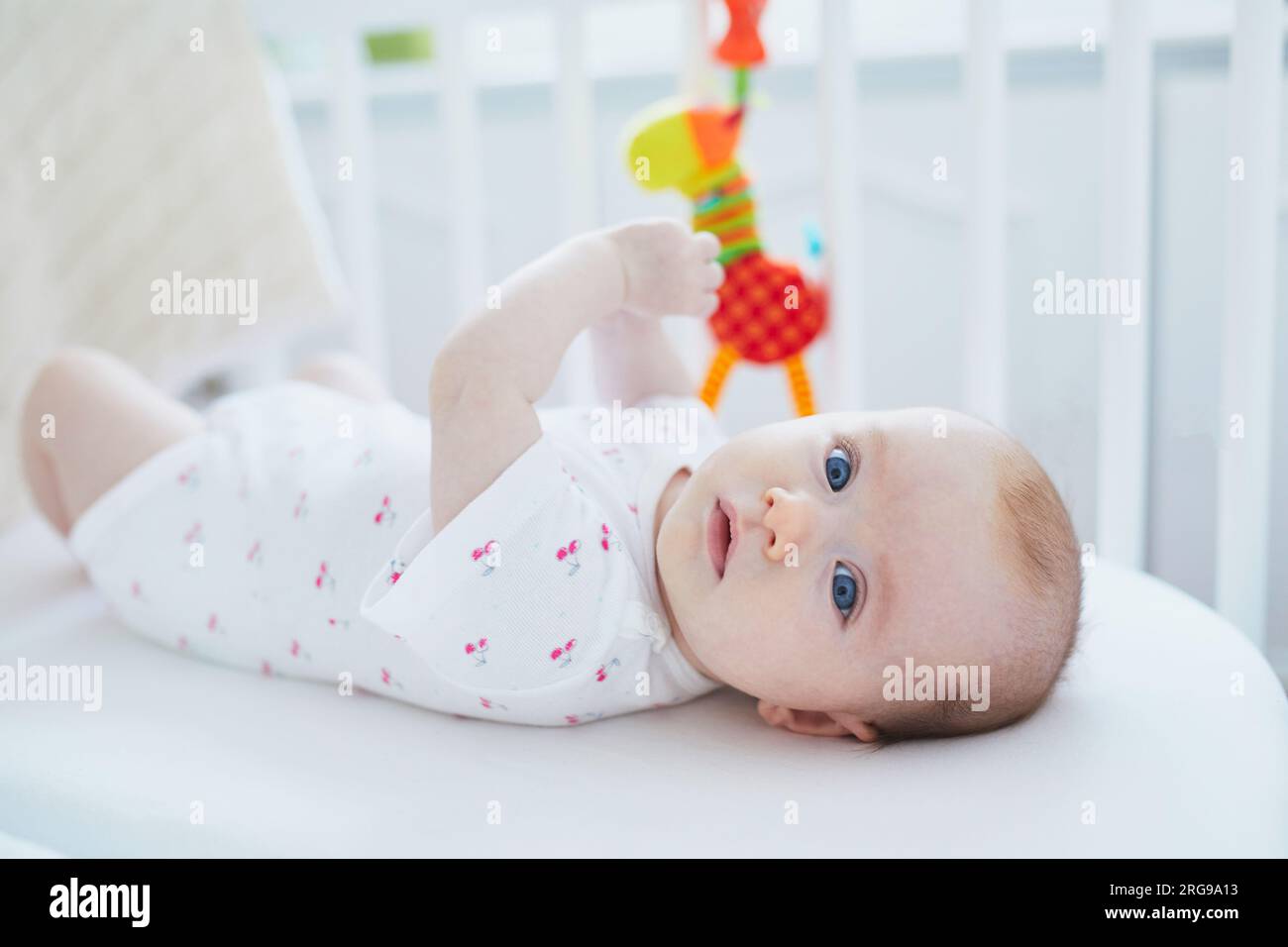 Baby girl lying in co-sleeper attached to parents' bed and playing with a toy Stock Photo