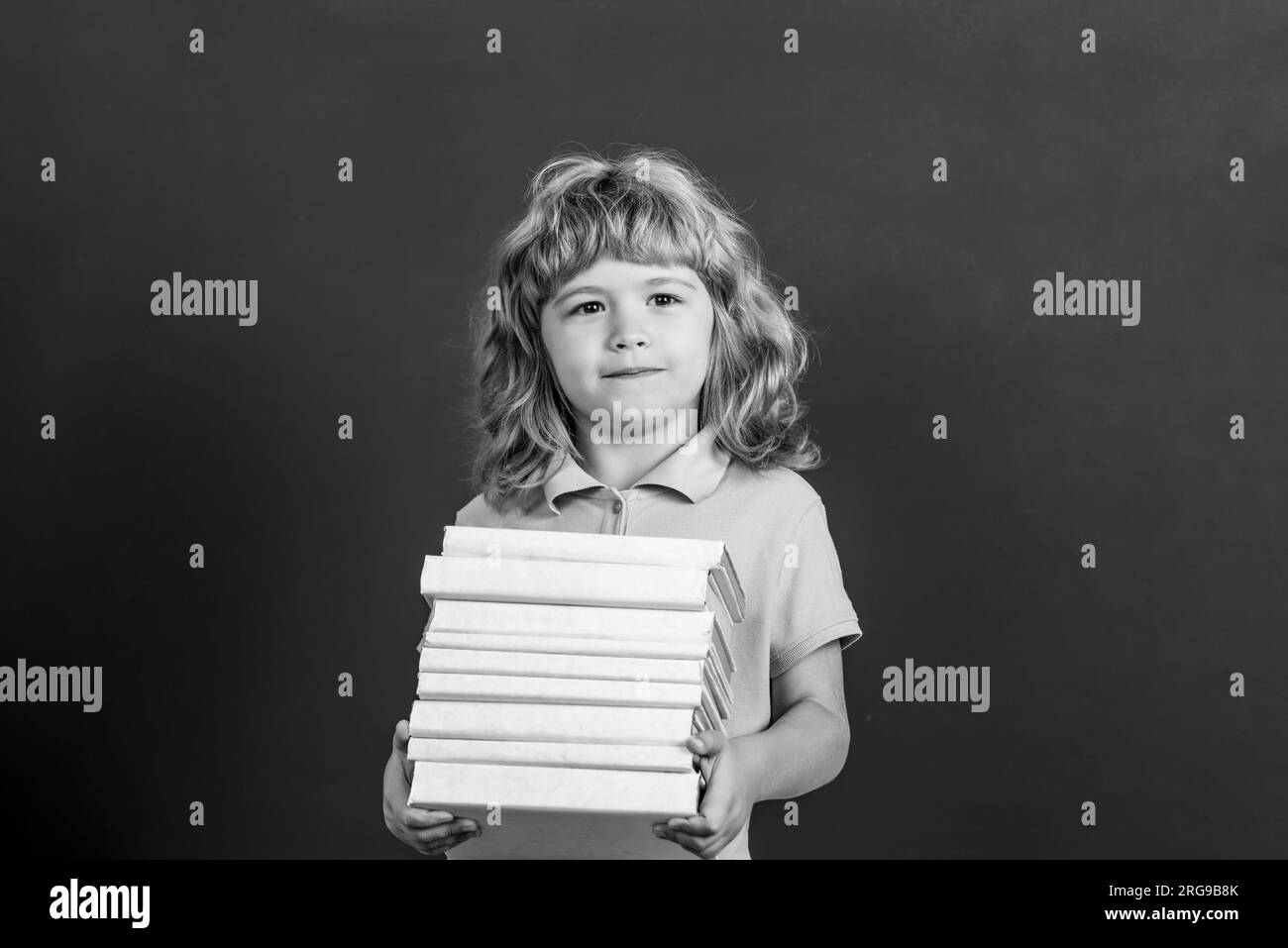 Education and creativity concept. Child holding stack of books with mortar board on blackboard. Back to school. Schoolchild in class. Black and white Stock Photo