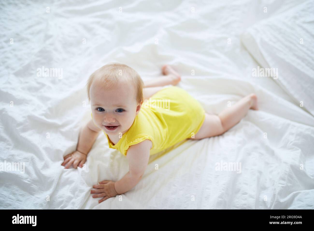 Baby girl crawling on bed and laughing. Happy healthy little child in yellow clothes. Infant kid in sunny nursery Stock Photo