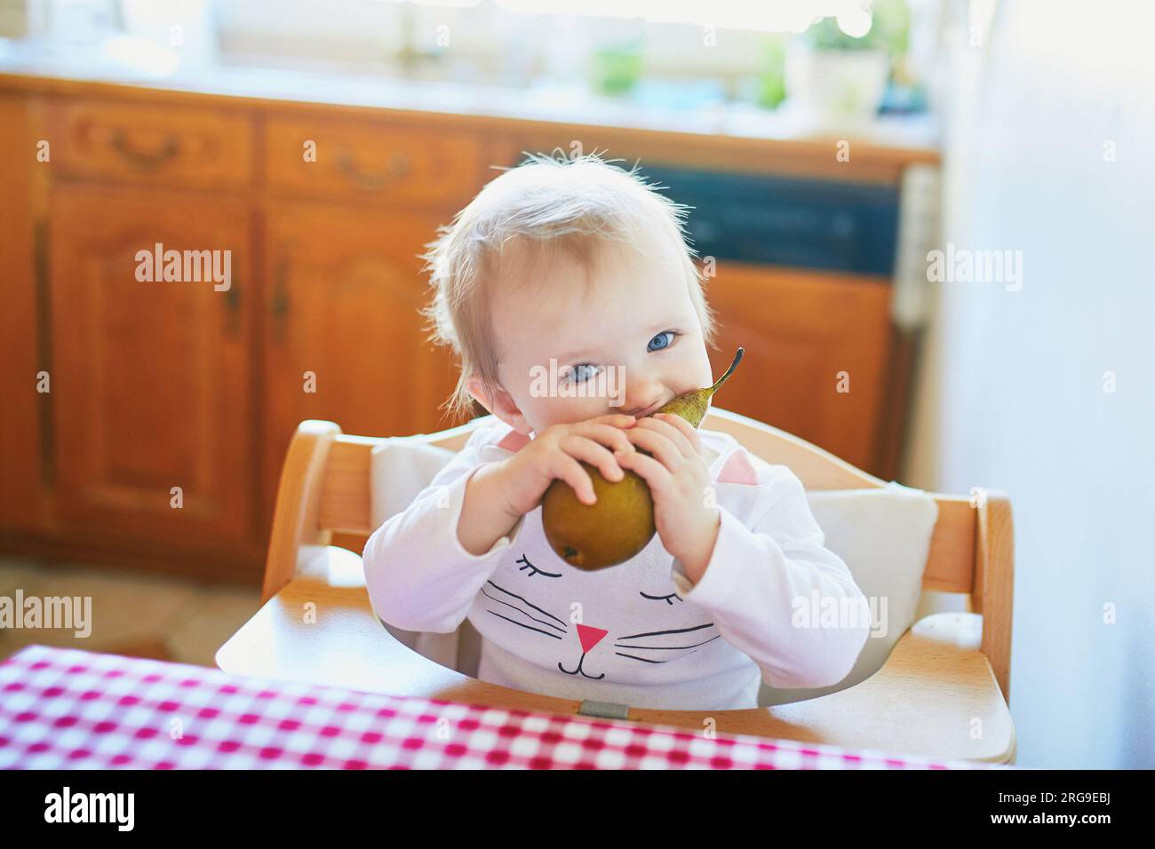 Cute baby girl eating pear in the kitchen. Little kid tasting solids at