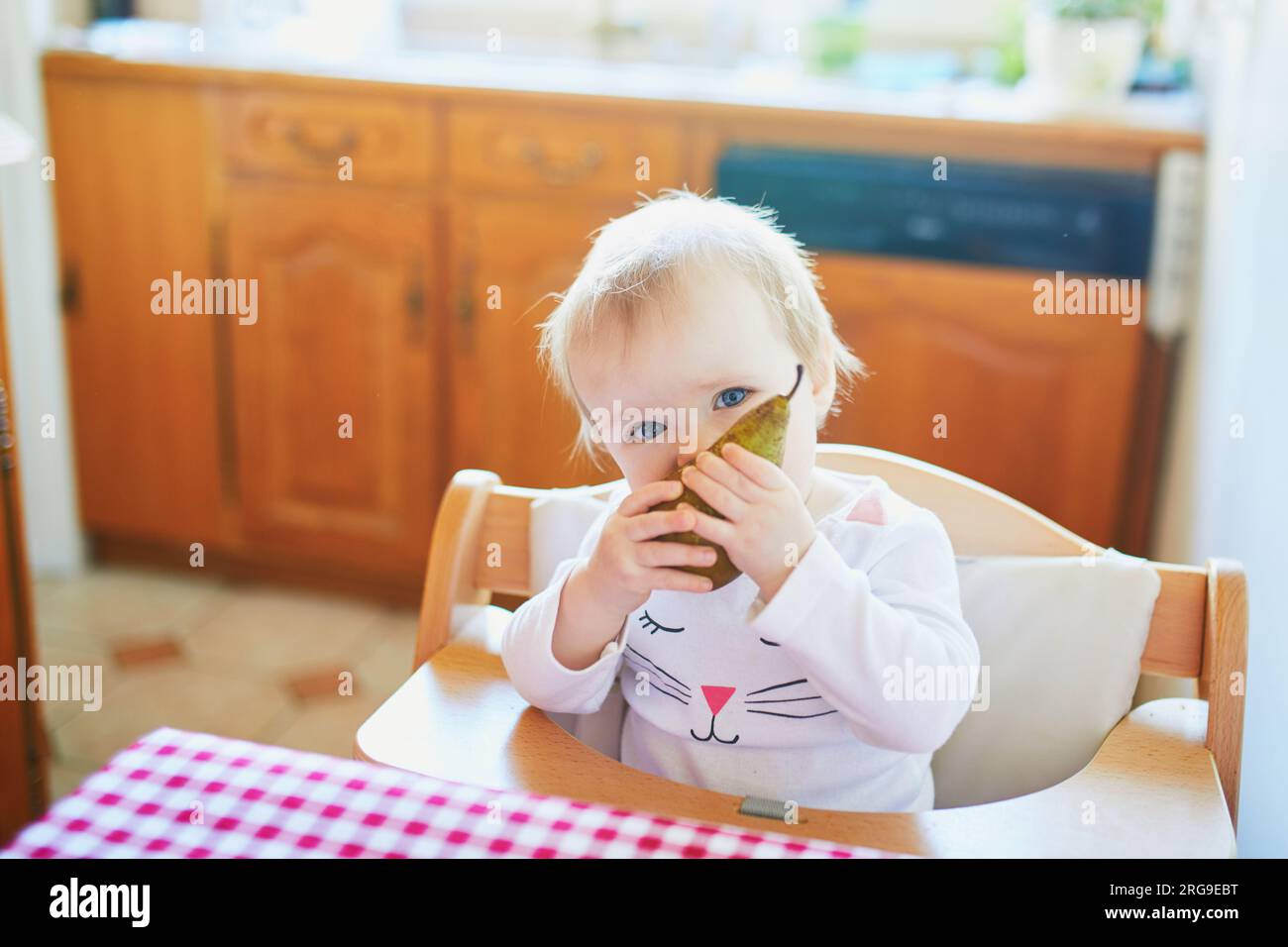 Cute baby girl eating pear in the kitchen. Little kid tasting solids at