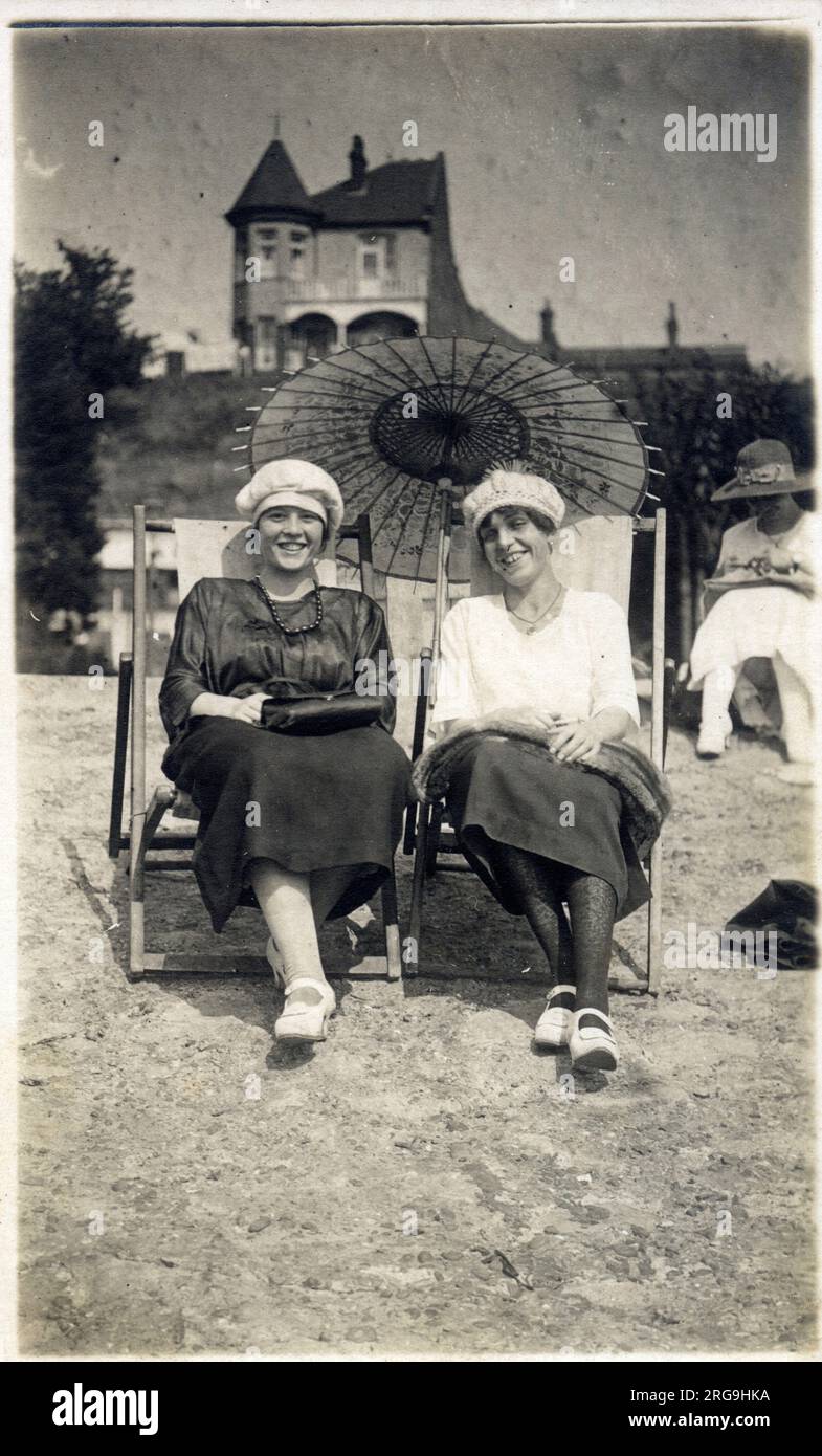 A stylish jovial pair of ladies sat in deckchairs on the beach beneath a pretty oriental paper parasol. Stock Photo