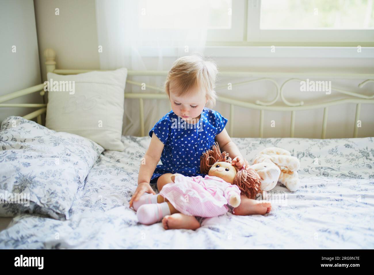 Adorable baby girl in blue dress sitting on bed and playing with a doll ...