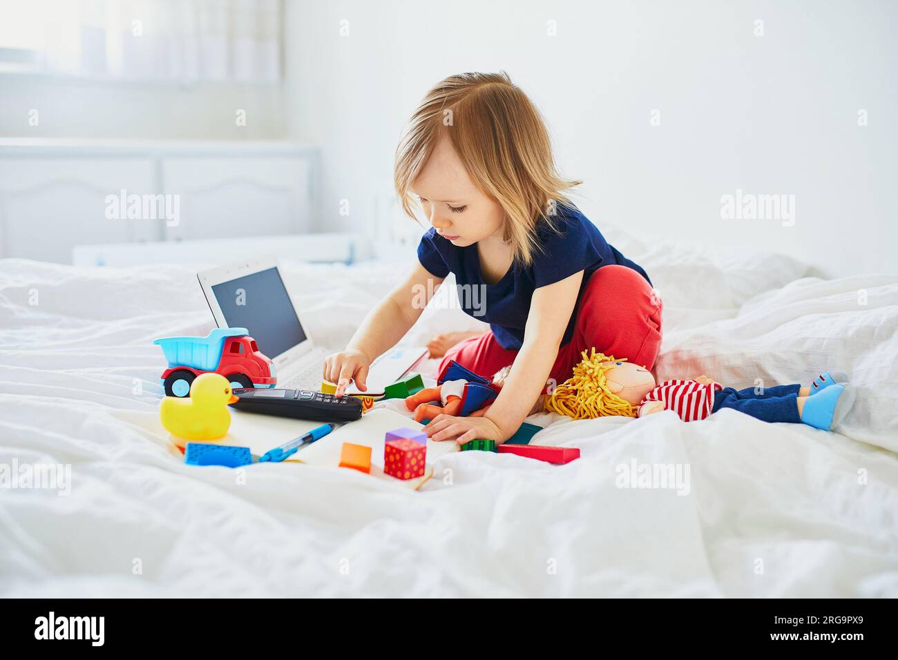 Toddler girl with laptop, notebook, phone and different toys in bed on clean white linens. Freelance, distance learning or work from home with kids co Stock Photo