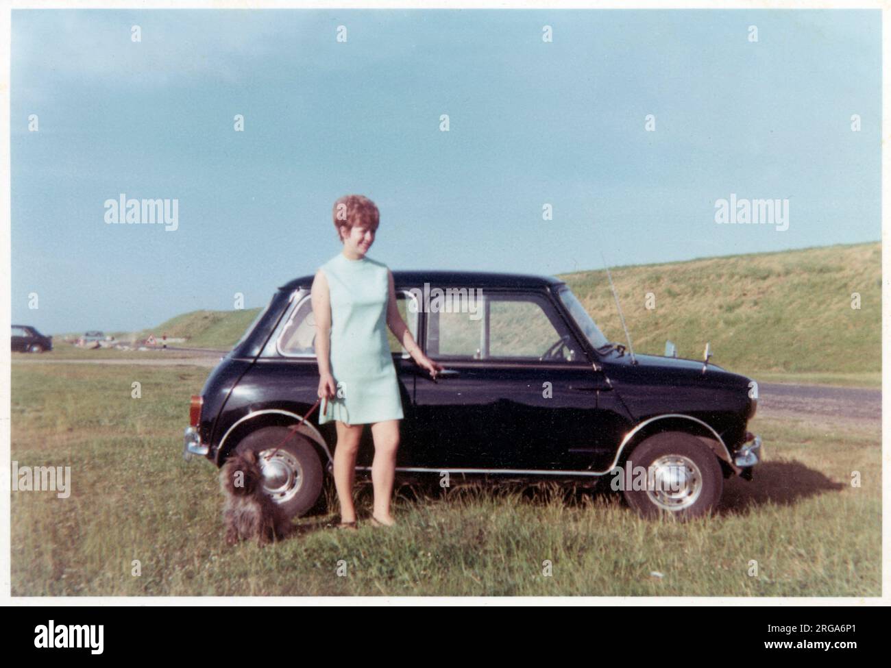 Young lady (and terrier) standing proudly next to her black Mini Cooper, parked on top of a coastal cliff - circa 1970. Stock Photo