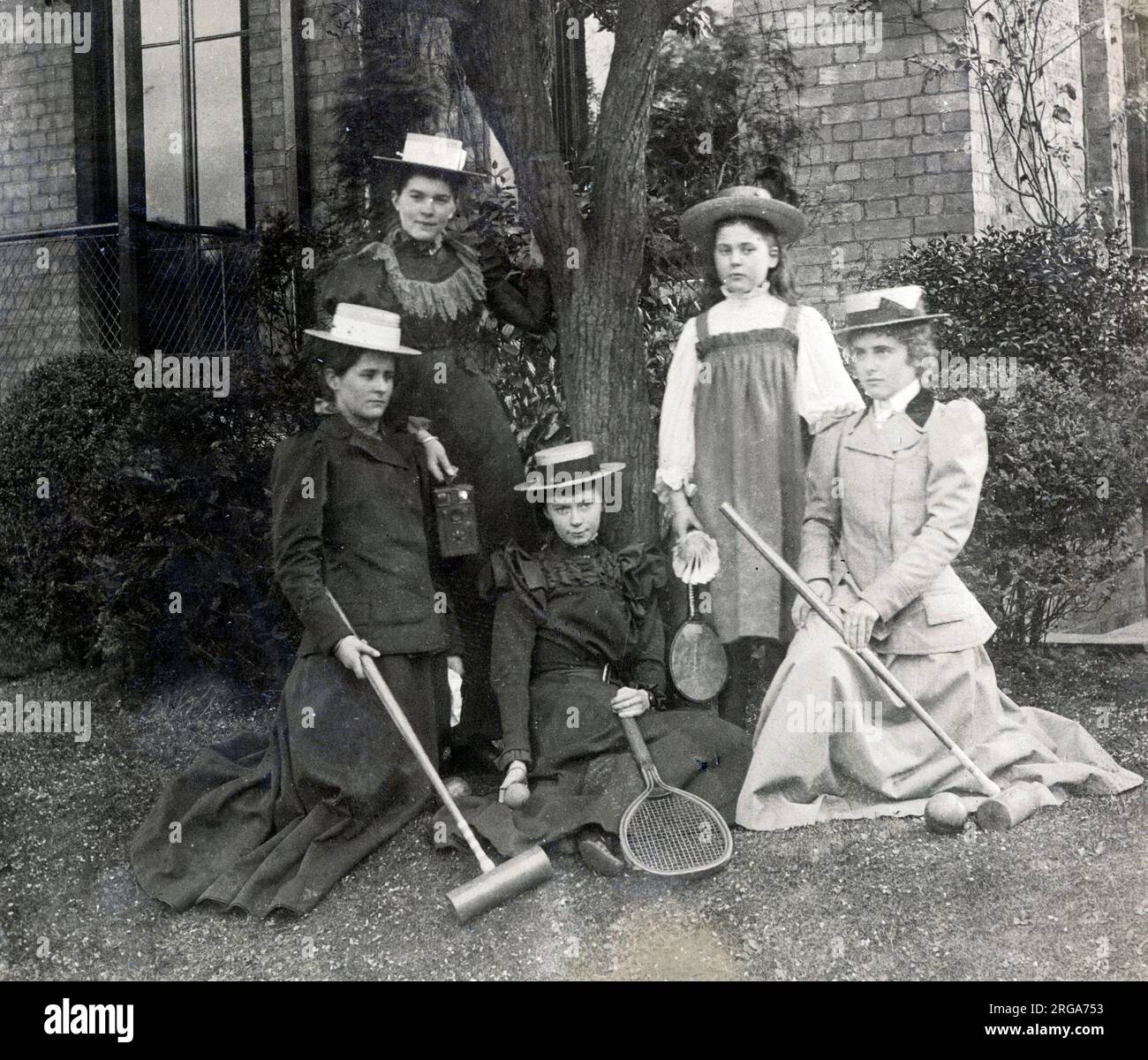 A group of women and girls, probably from the same family, pose for a photograph in the garden with various sporting accoutrements - croquet mallets, tennis racquets and badminton racquet and shuttlecock - all popular pursuits for the late Victorian or Edwardian lady. The girl back left is holding a photographic camera. Stock Photo