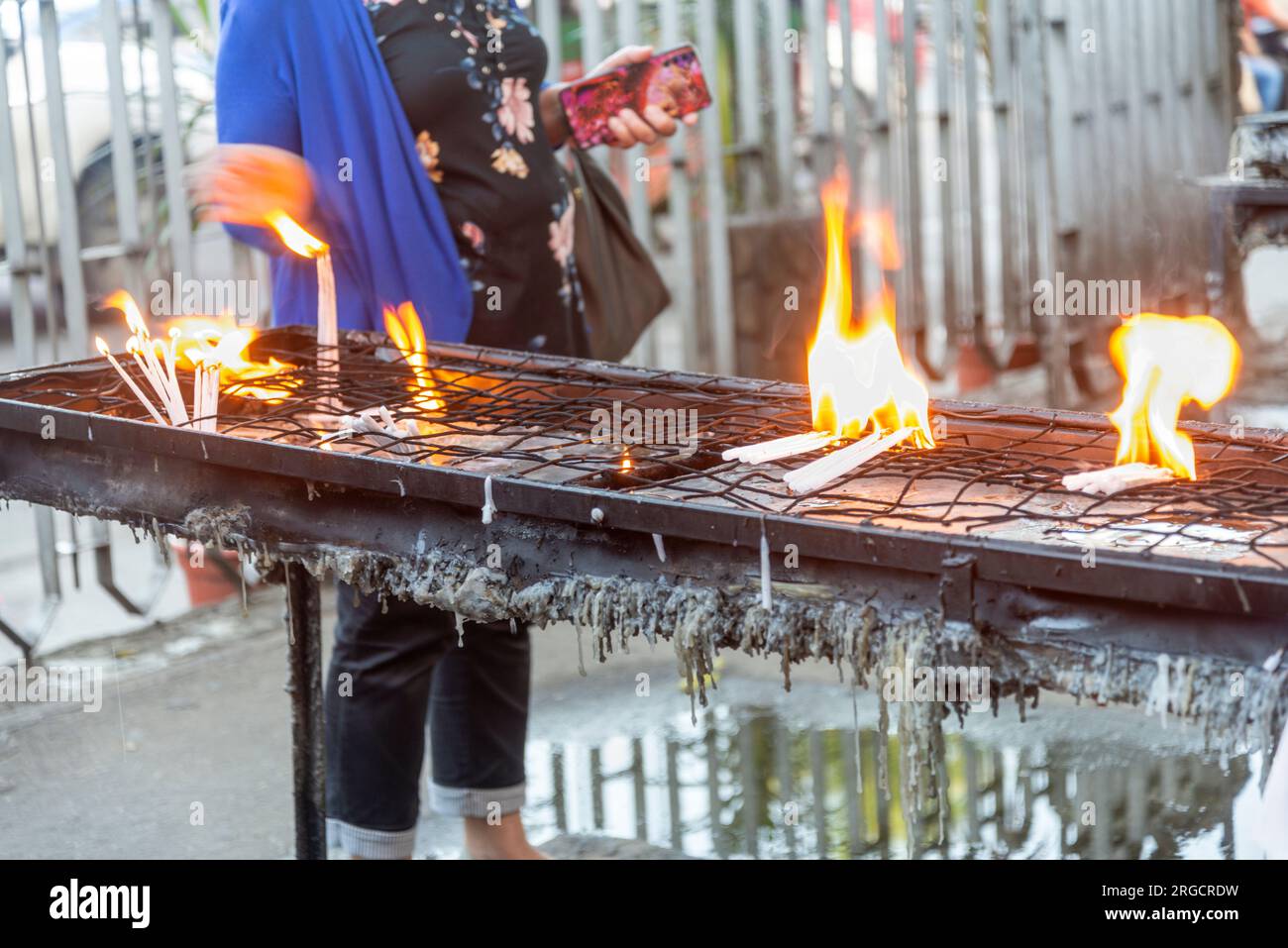 Large bright flames from burning candles,molten candle wax dripping from metal tray as people place and position them carefully as they pray to Jesus Stock Photo