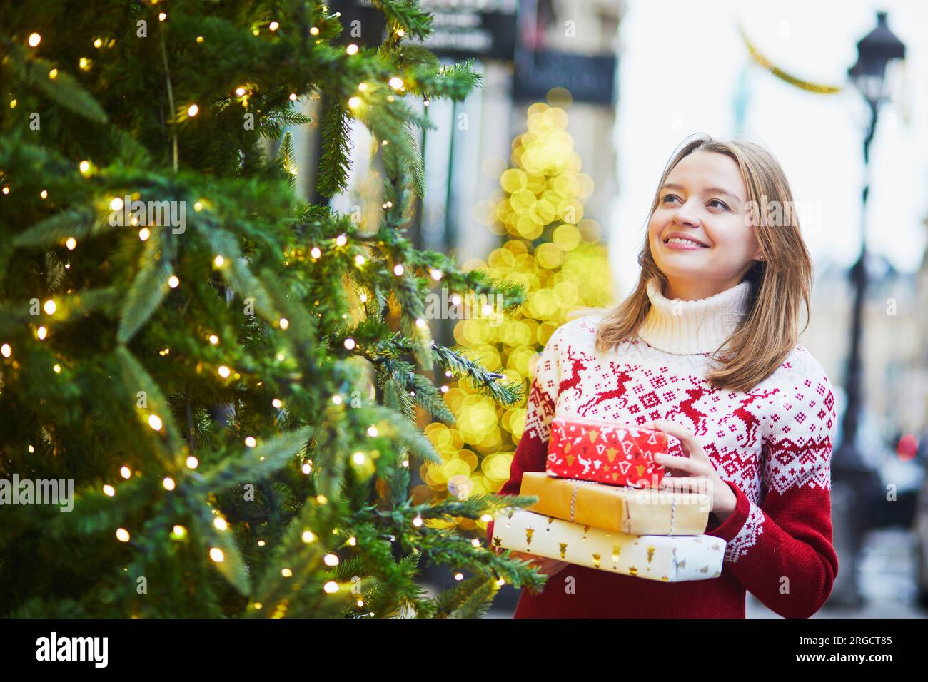 Happy young girl in warm red knitted sweater with pile of holiday gifts on a street of Paris decorated for Christmas Stock Photo