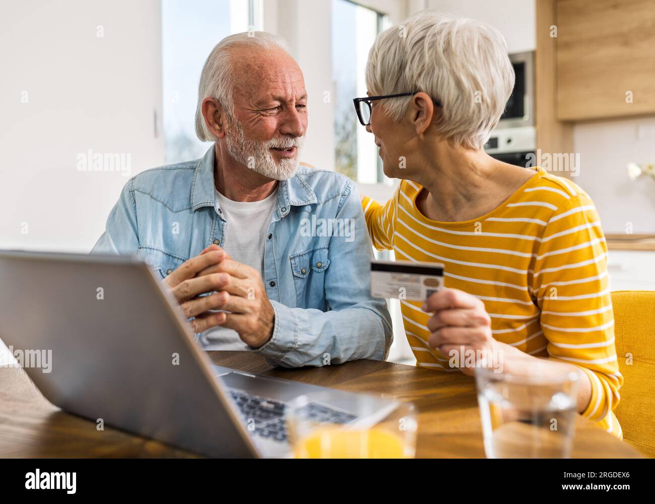 Cheerful senior couple sitting at dining table with kitchen in background and purchasing online with credit card Stock Photo