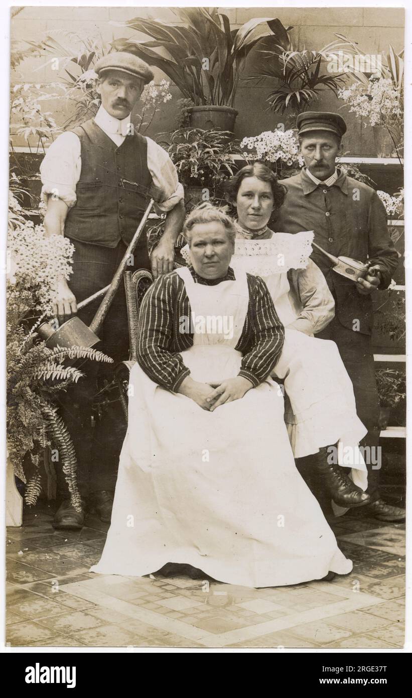 Four servants pose for a photograph in what appears to be a conservatory. The gardener, with long-spouted watering can, is on the left, and a chauffeur and mechanic, with an oil can, on the right, stand either side of two women, possibly a cook, or housekeeper and a maid. Stock Photo