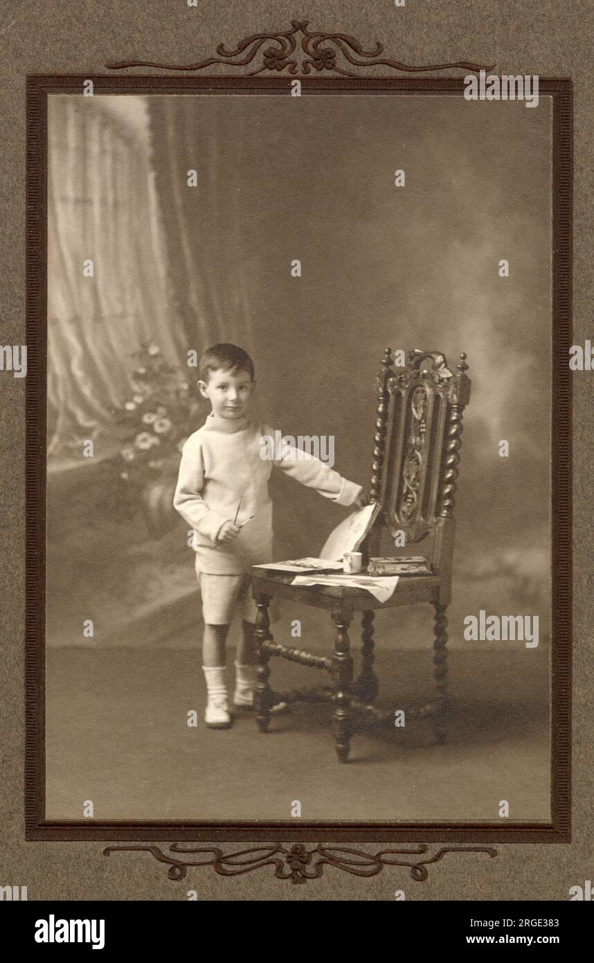 Charming studio photograph of a small boy posing with paintbrushes in hand and paints and painting books laid out on a chair. Stock Photo