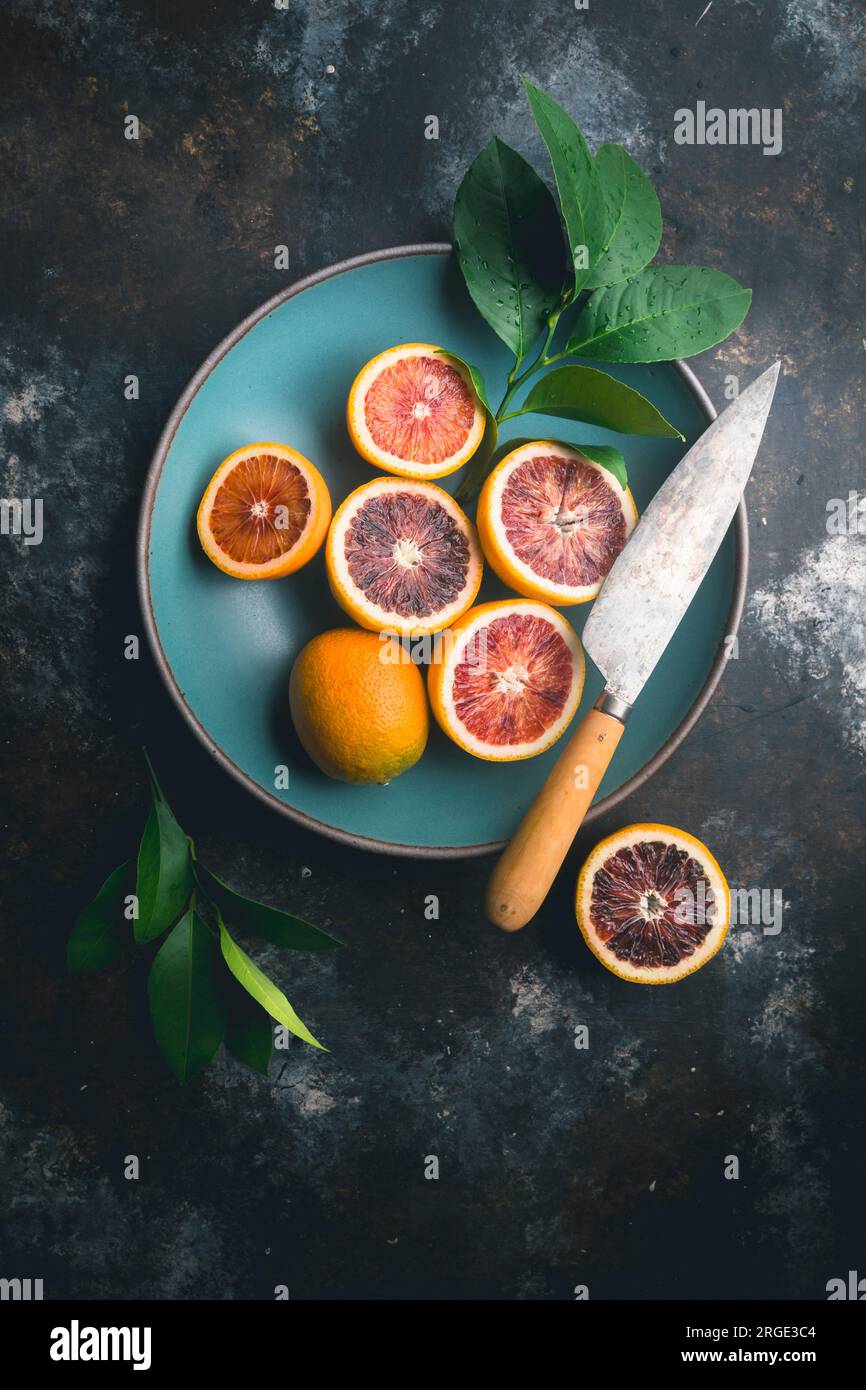 Blood Oranges, halved and whole with citrus leaves, in blue ceramic bowl and on table, with knife on dark background Stock Photo
