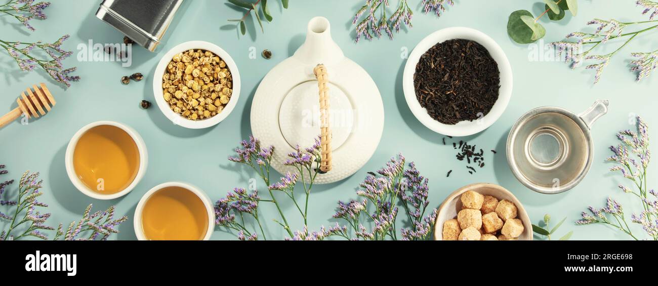 A white porcelain teapot, two cups, suga and assortment of dry tea in white ceramic bowls on blue background.  Top view. Banner Stock Photo