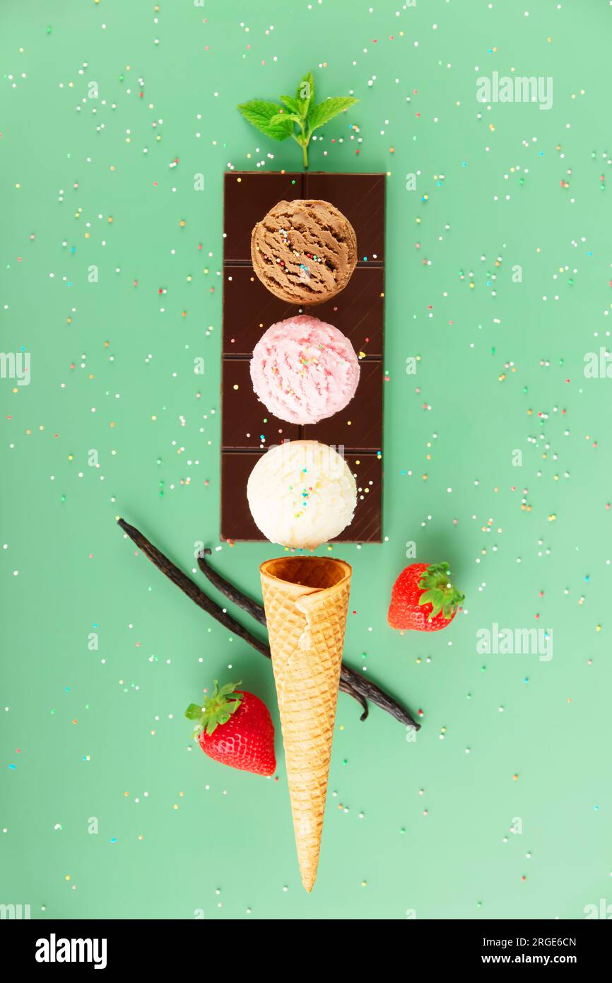 Chocolate, vanilla, strawberry scoops, waffle cone and ingredients on green background with colorful sprinkles, levitating concept. Spring or summer m Stock Photo