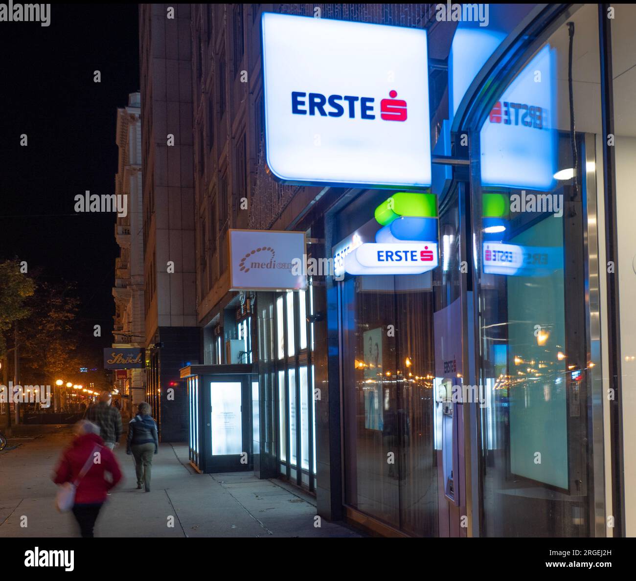 Pedestrians walk past the illuminated shop window of Erste Bank at night Stock Photo