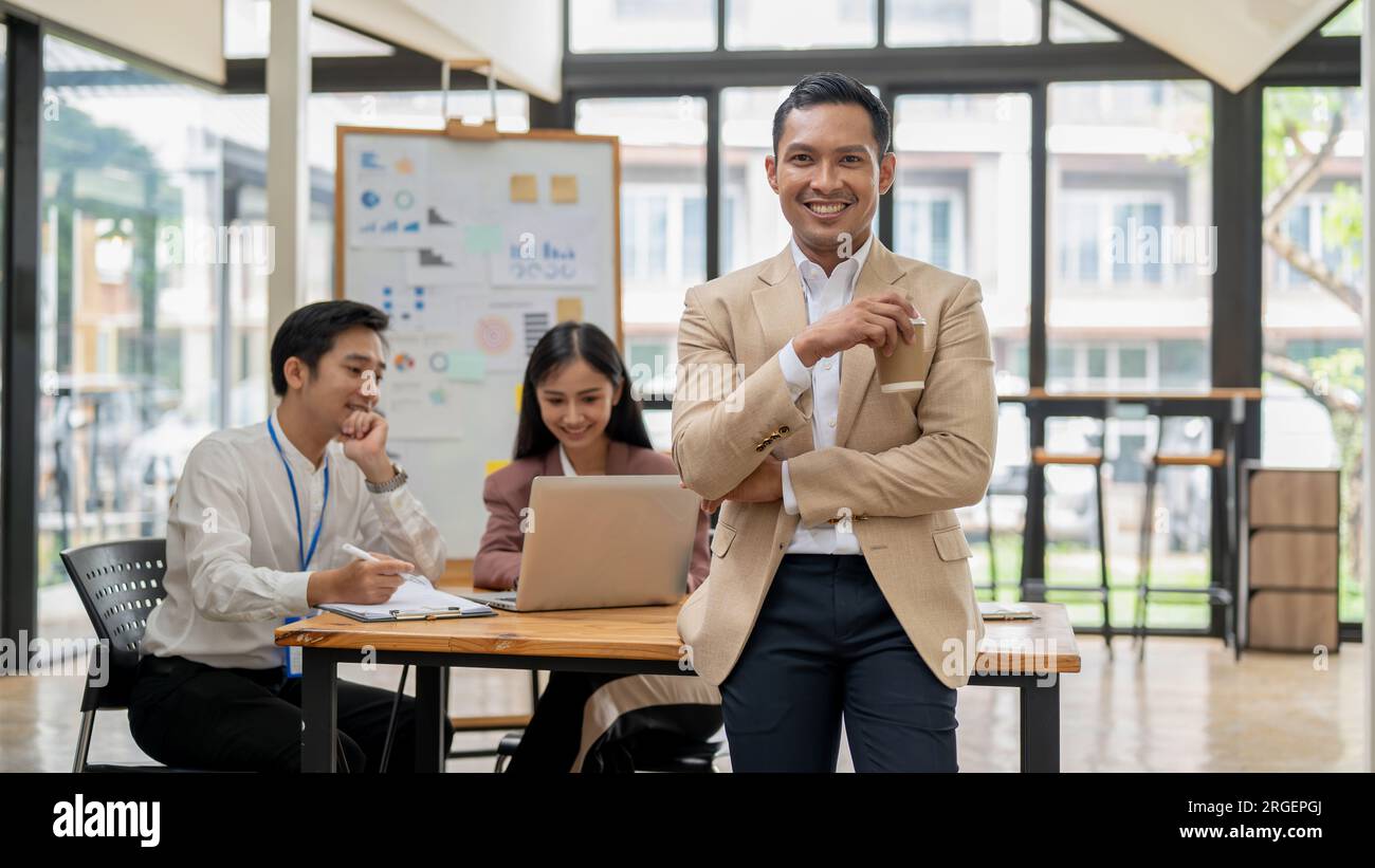 A handsome and successful Asian male CEO or businessman stands with a coffee cup in his hand, smiling at the camera, while his employees are working a Stock Photo