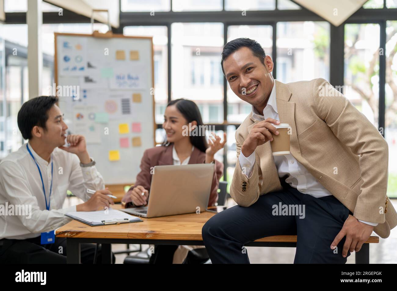 A handsome and successful Asian male CEO or businessman sits on a table with a coffee cup in his hand, smiling at the camera, while his employees are Stock Photo