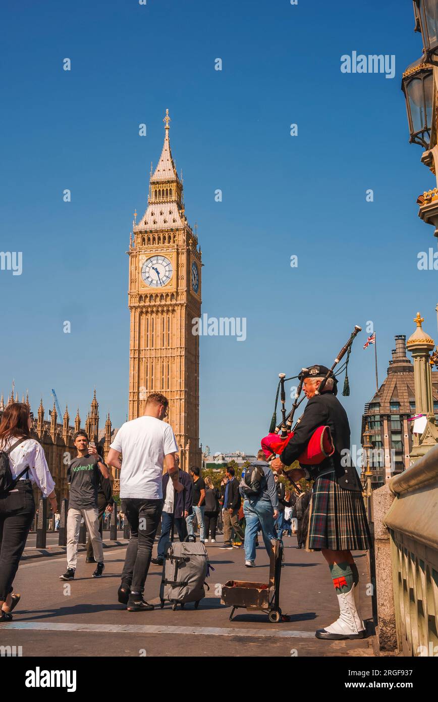 Senior man playing bagpipe amidst Tourists walking by Clock Tower Stock Photo