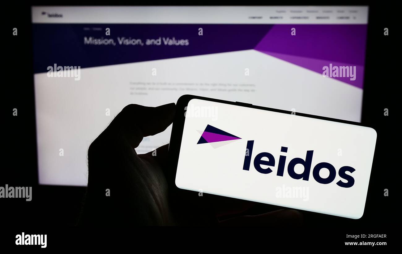 Person holding cellphone with logo of US defense company Leidos Inc. on screen in front of business webpage. Focus on phone display. Stock Photo
