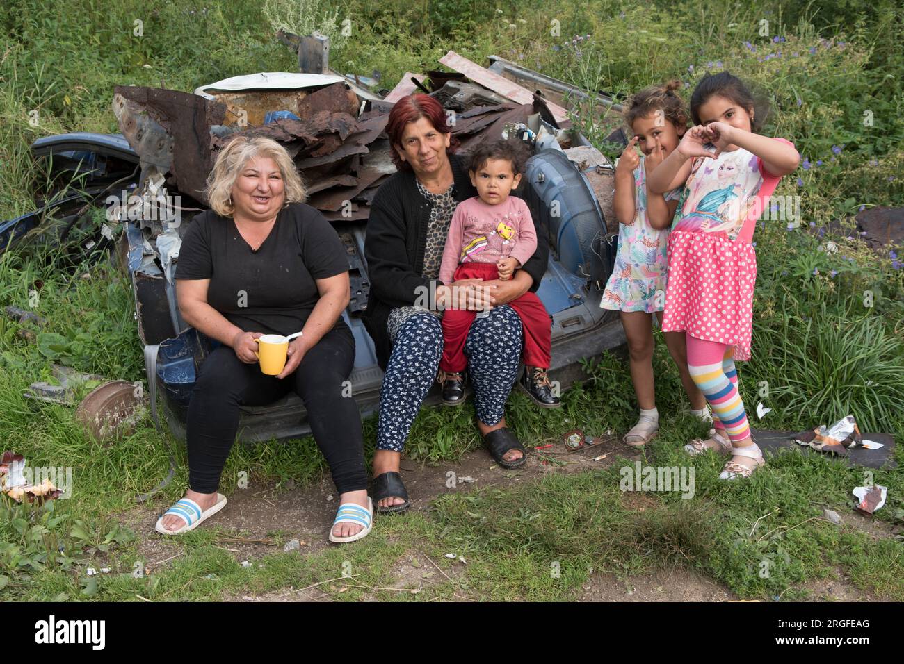 Romany gypsy grandmothers with grandchildren. They are sitting on a scrapped car across the road from their homes. The two girls are making with their hands a heart sign and seeing sign - friendship. Gypsy settlement on the outskirts of Sumiac, Brezno District, Slovakia August 2023 2020s HOMER SYKES Stock Photo