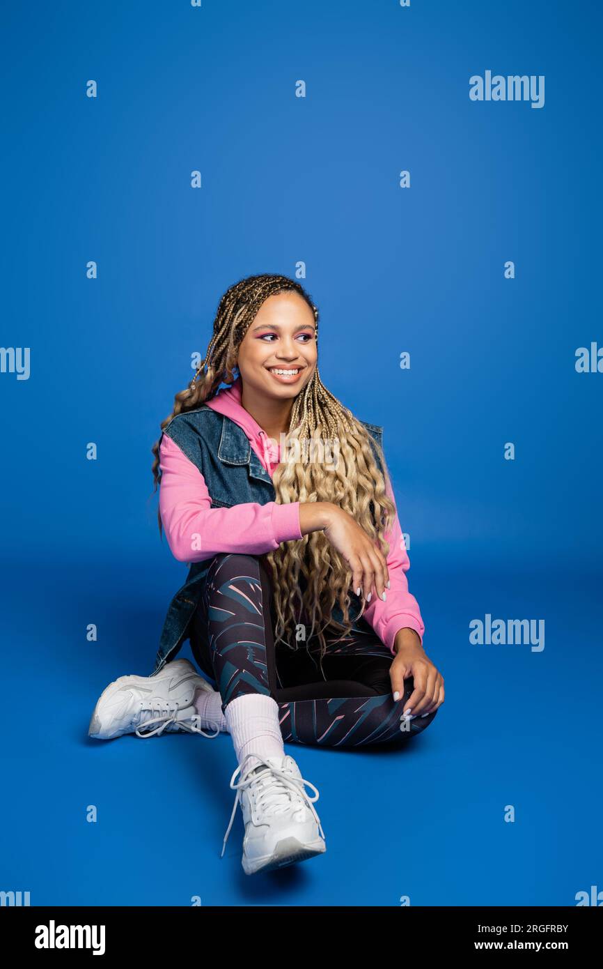 full length of young long haired african american woman sitting in sporty outfit on blue background Stock Photo