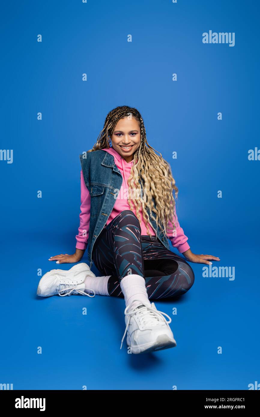 full length of joyful long haired african american woman sitting in sporty outfit on blue background Stock Photo