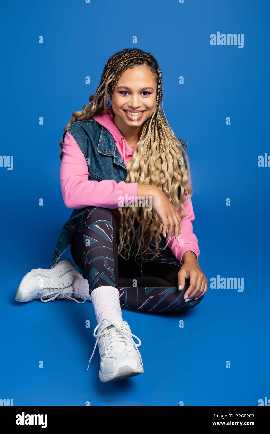 full length of happy long haired african american woman sitting in sporty outfit on blue background Stock Photo