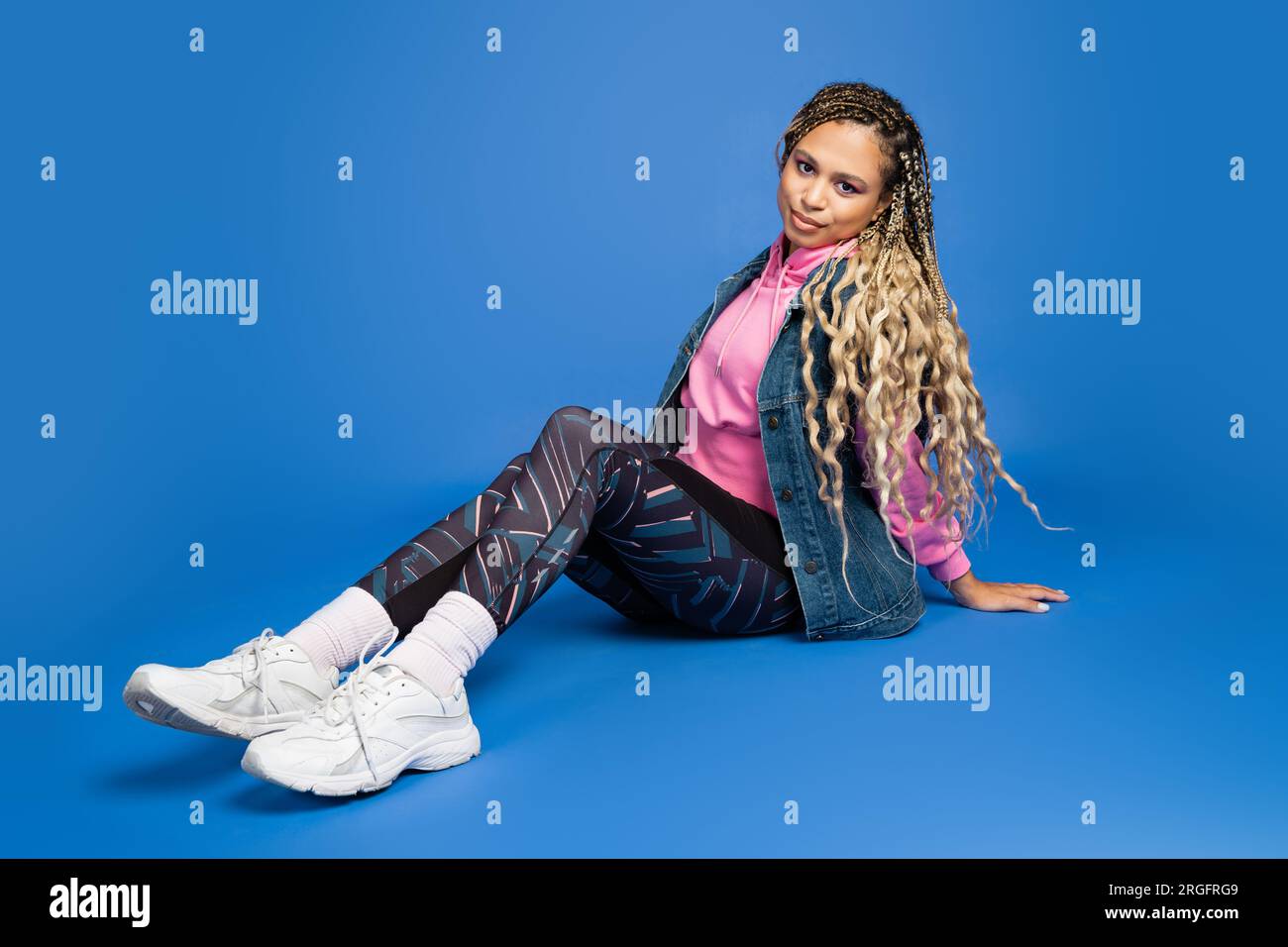 full length of long haired african american woman in sporty outfit sitting on blue background Stock Photo