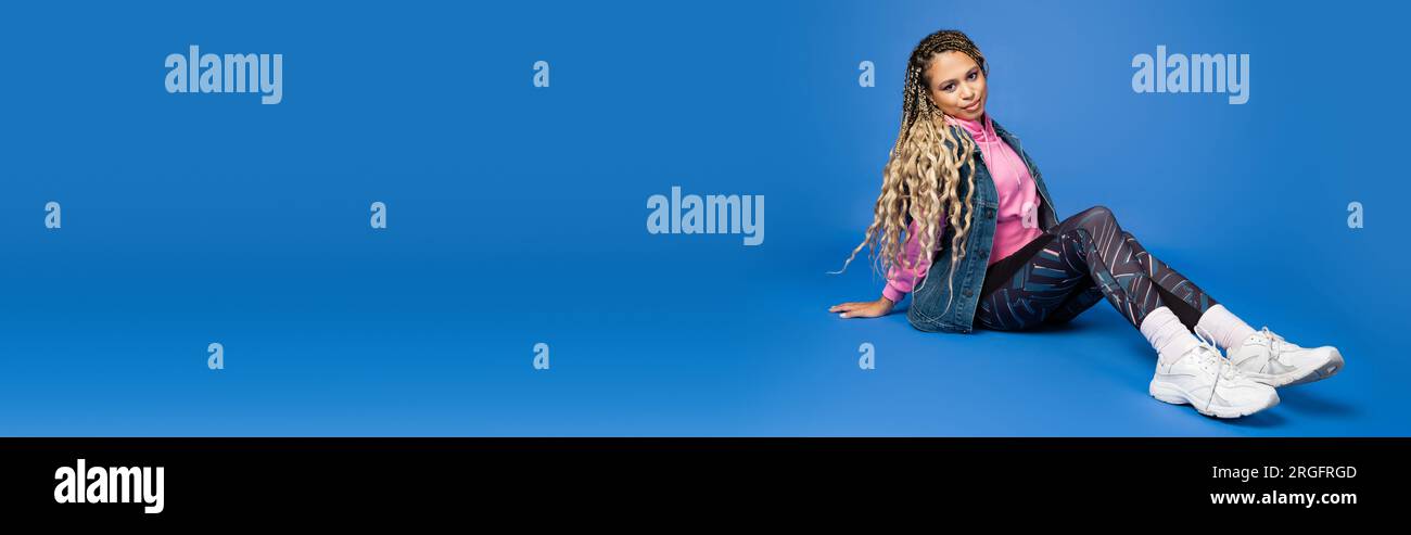 full length of long haired african american woman in sporty outfit sitting on blue, banner Stock Photo