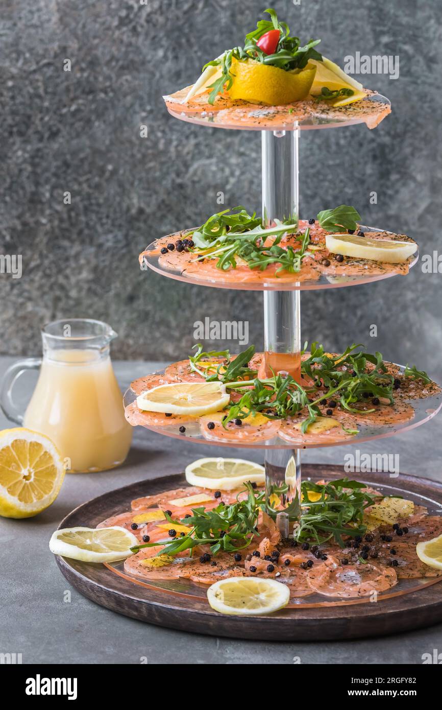 Salmon carpaccio with lemon and rocket on a cake stand, italian starter, antipasti, gray background, vertical. Stock Photo