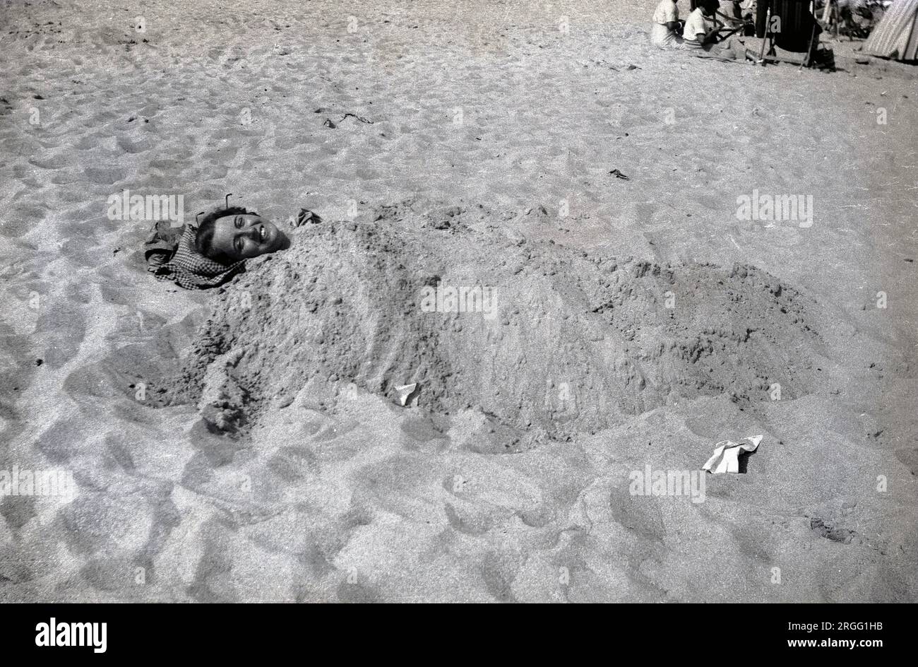 1940s, historical, summertime and a lady lying on a beach, with her body and feet covered in sand, with a big smile on her face. Stock Photo