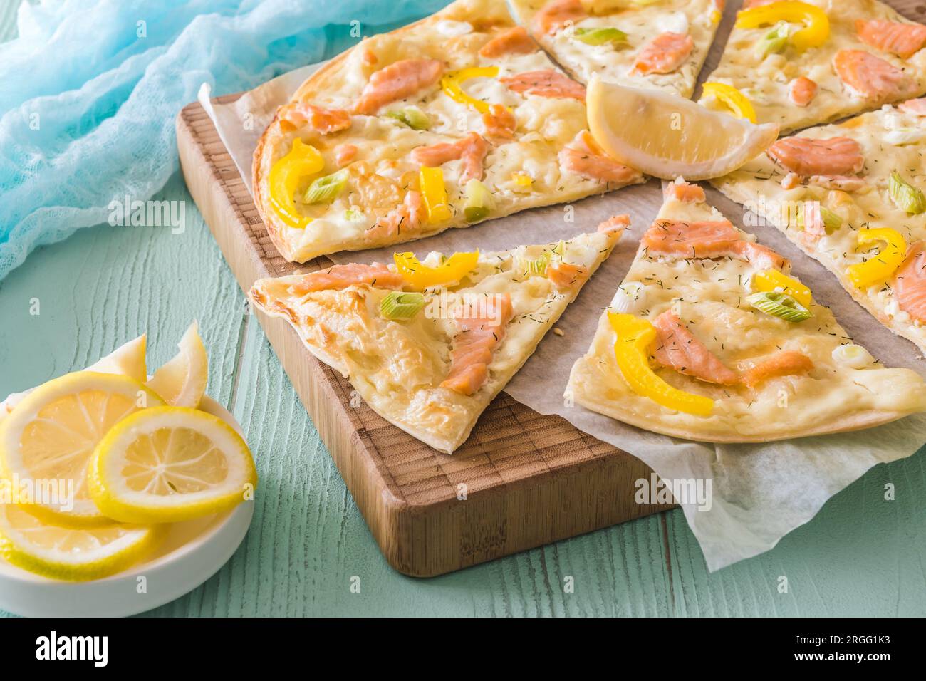 Pieces of tarte flambee with salmon, yellow pepper and spring onions on a wooden board on light green background Stock Photo