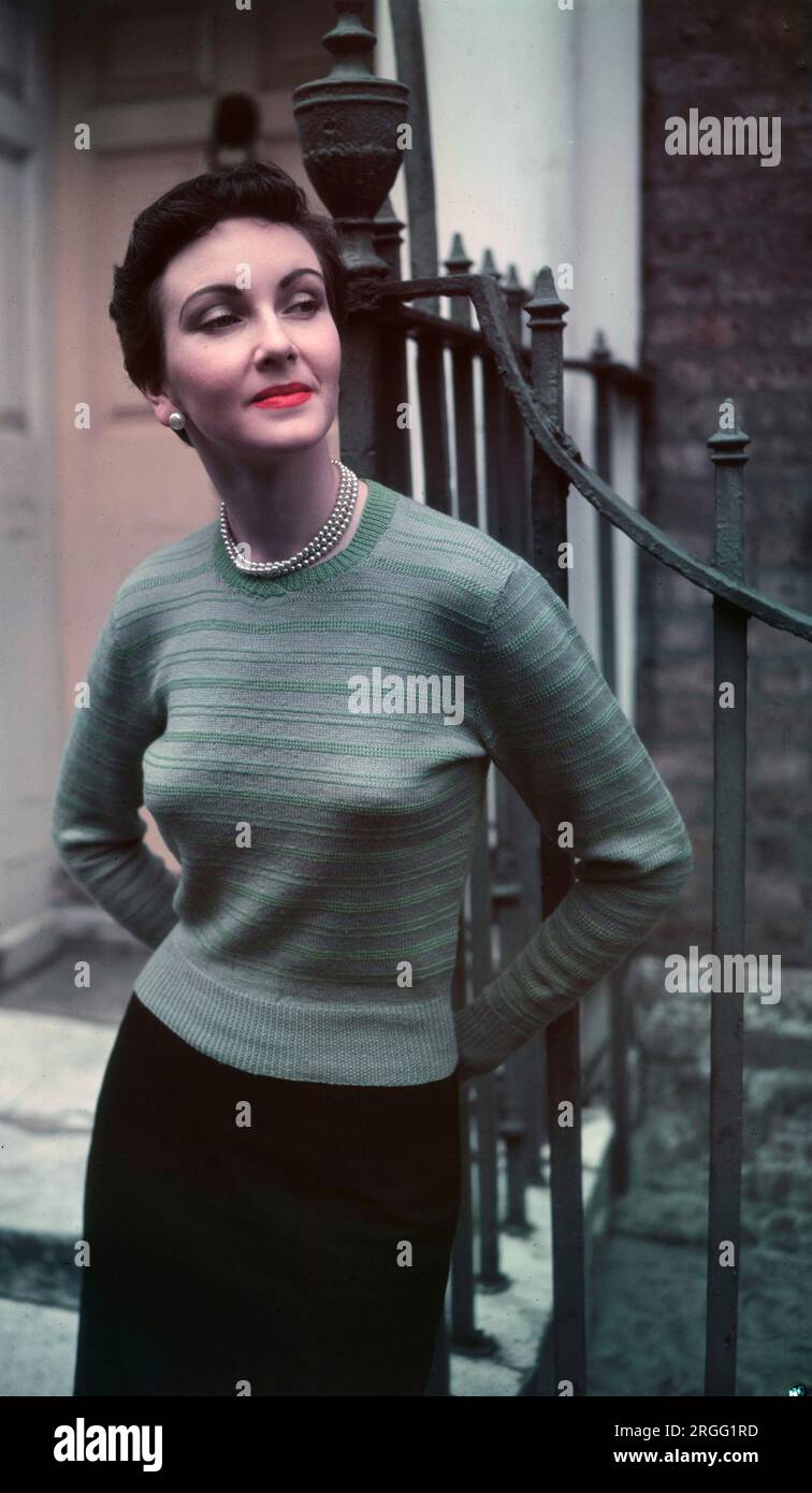 1950s, historical, standing outside by iron railings, an elegant, attractive lady wearing a light ribbed woollen sweater and pencil skirt, clothes that reflect her feminine, delicate features and refined, sophisticated style, England, UK. Woollen tops and sweaters, worn over a cone-or bullet-shaped bra as seen here, were a popular female clothing item in this era, as new yarns and colours were introduced. Stock Photo