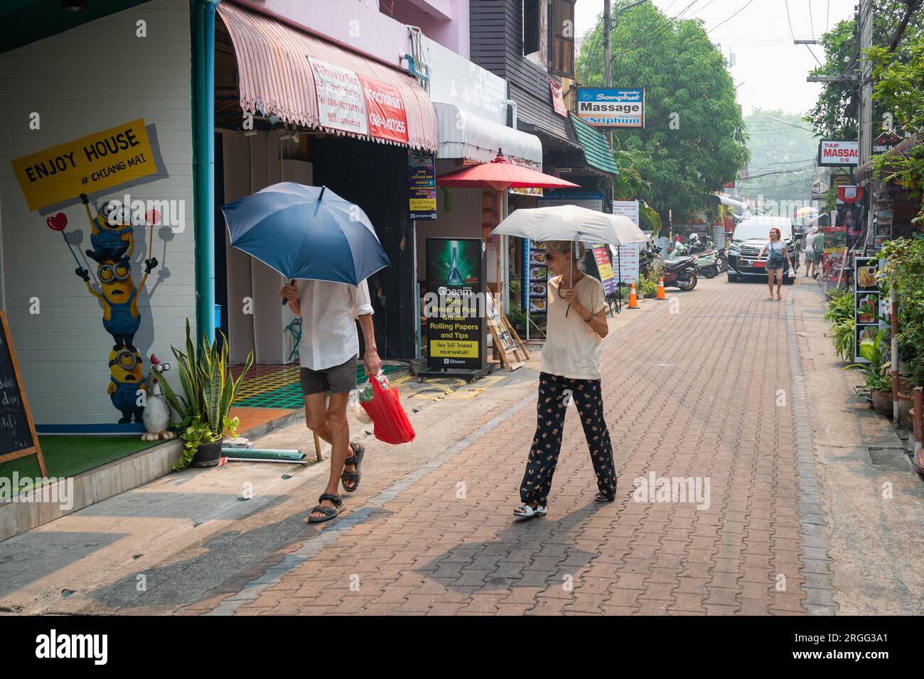 Couple of senior, stylish tourists with sun umbrellas walk down Moonmuang Road in Chiang Mai Stock Photo