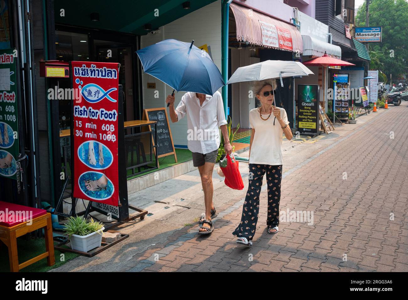 Couple of senior, stylish tourists with sun umbrellas walk down Moonmuang Road in Chiang Mai Stock Photo