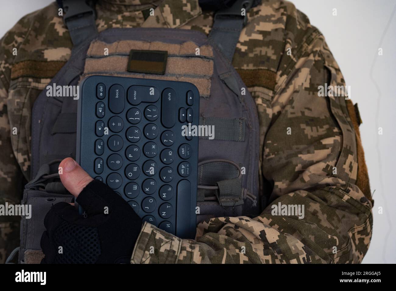 Ukrainian soldier in grey bulletproof jacket with icon of ukrainian flag and keyboard in hand in black glove. Stock Photo