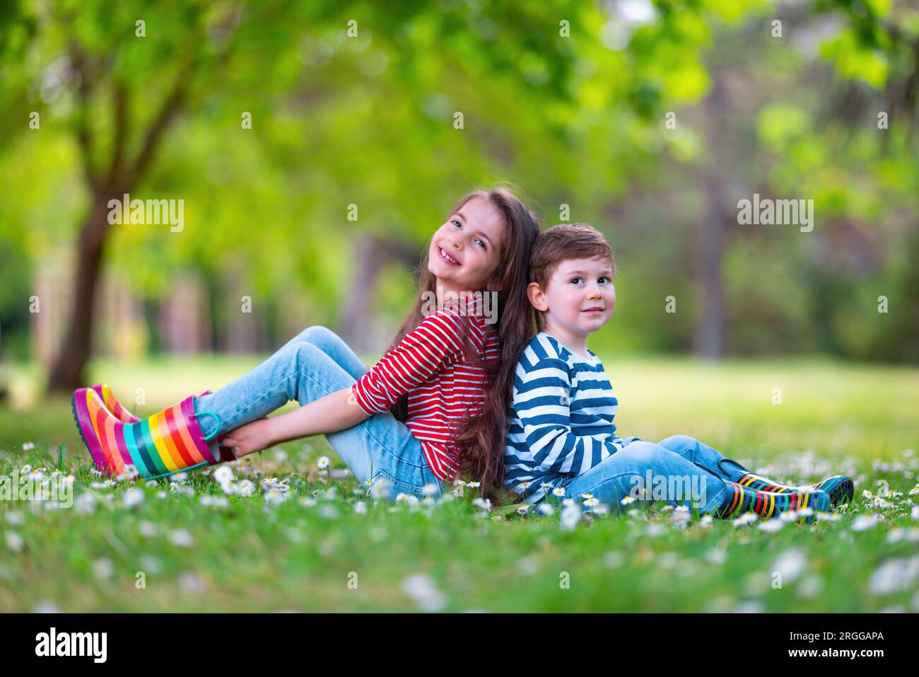 Happy kids boy and girl in rain rubber boots playing outside in the green park with blooming field of daisy flowers Stock Photo