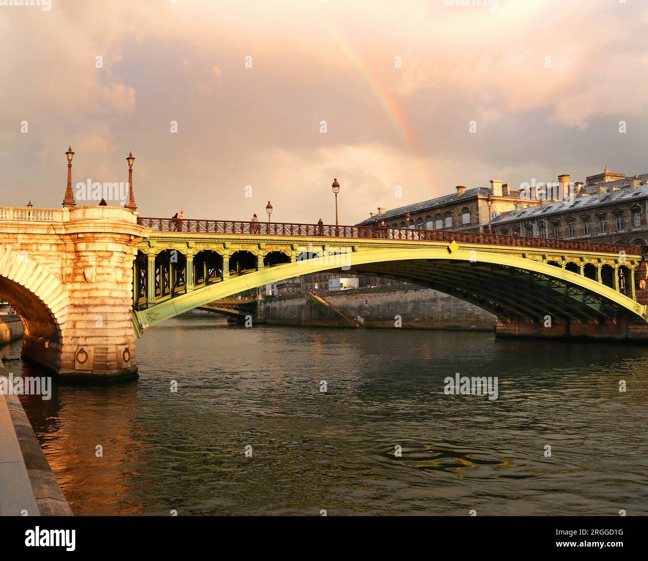 The green metal of the Pont Notre Dame, bridging the River Seine in Paris, glows in early evening golden light after heavy rain below a faint rainbow Stock Photo