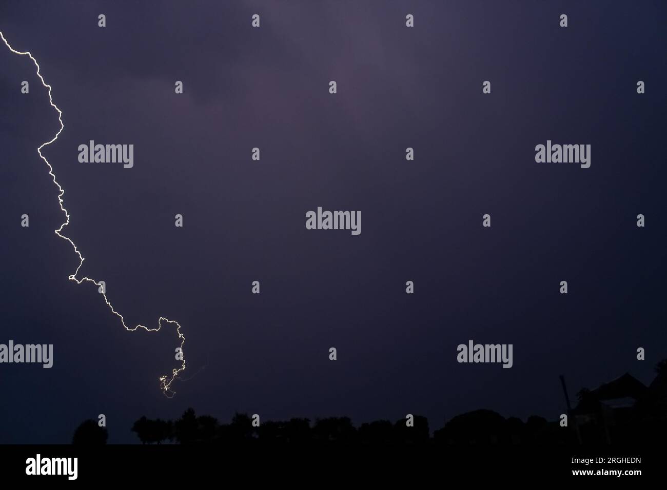 Rainy sky on a summer night with a flash of lightning on the left side of the sky, with the roofs of rural buildings. Stock Photo