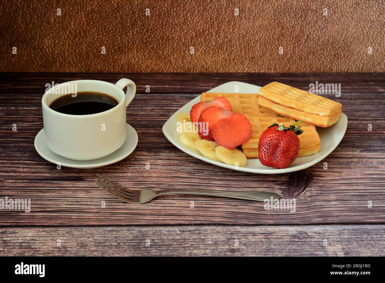 A cup of hot black coffee on a saucer and a plate with several strawberry and banana waffles on a wooden table. Close-up. Stock Photo