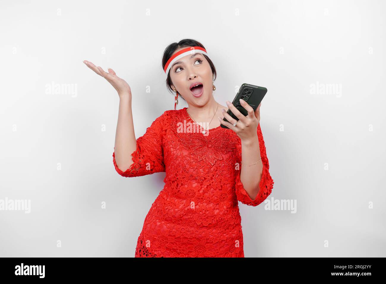 A happy Asian woman wearing red kebaya and headband, holding her phone, and pointing copy space on top of her, isolated by white background. Indonesia Stock Photo