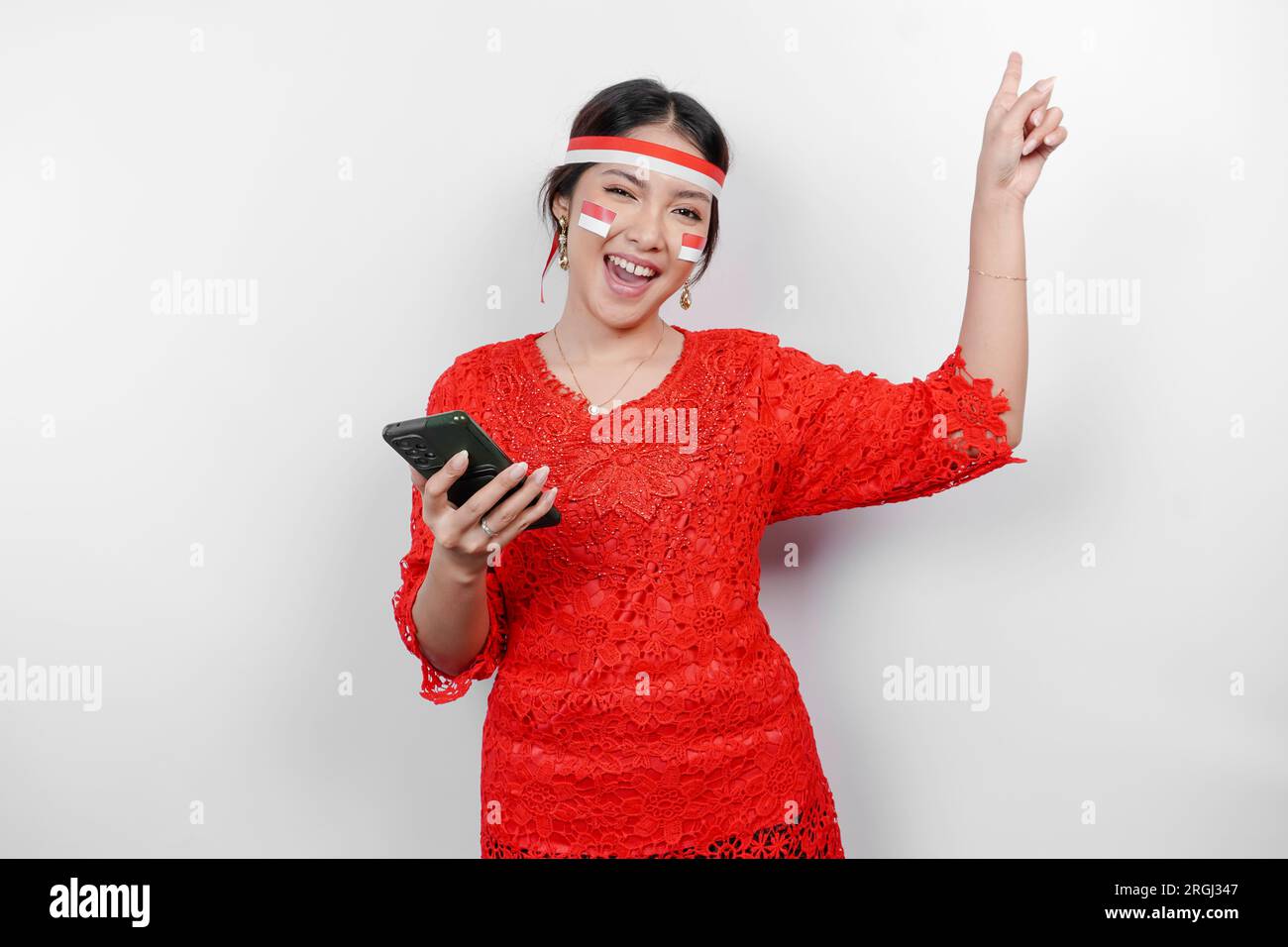 A happy Asian woman wearing red kebaya and headband, holding her phone, and pointing copy space on top of her, isolated by white background. Indonesia Stock Photo