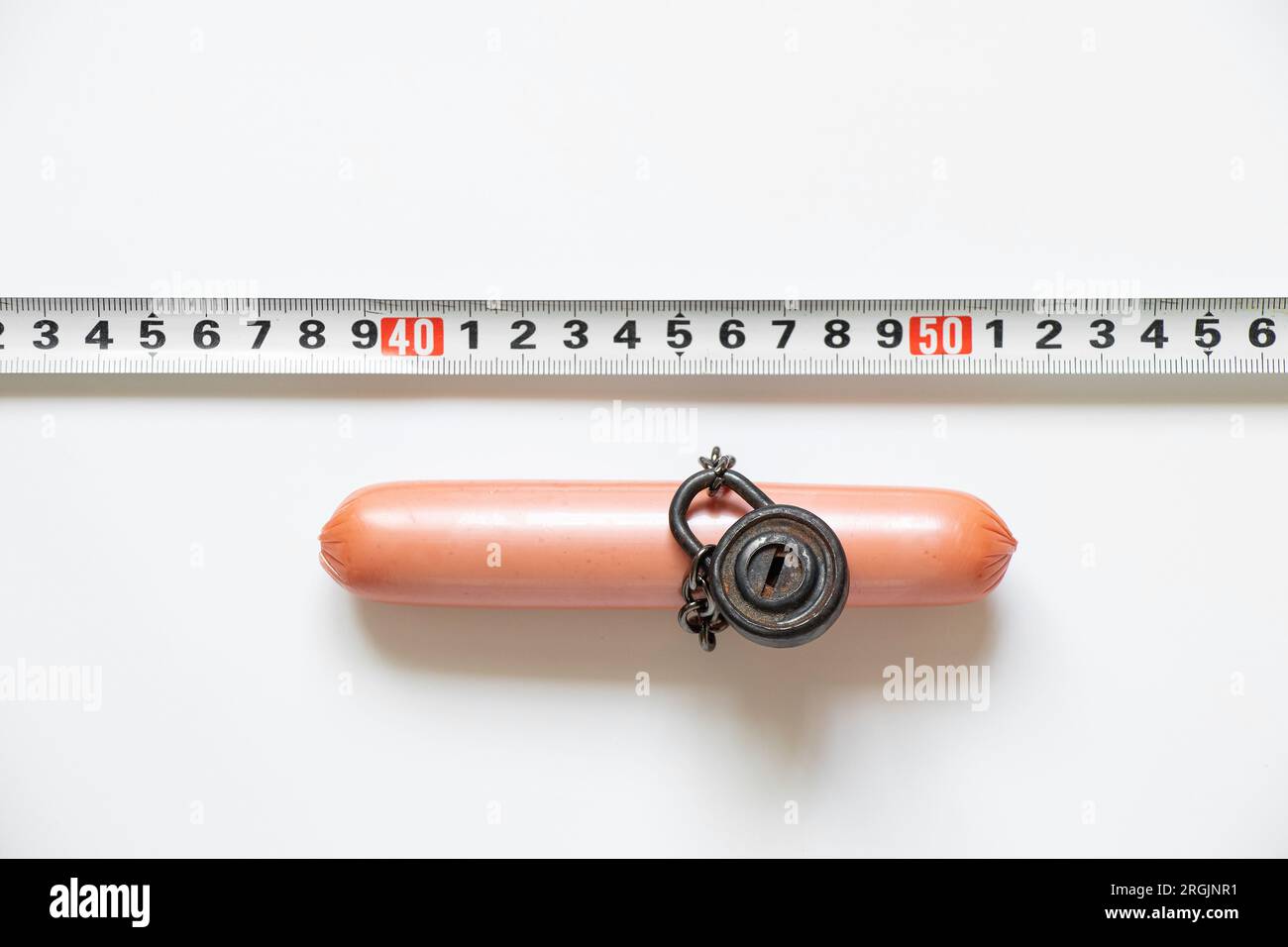 Sausage on a chain under lock and key and next to it lies a centimeter tape on a white background in the sun, rejection of unhealthy food, diet and ov Stock Photo