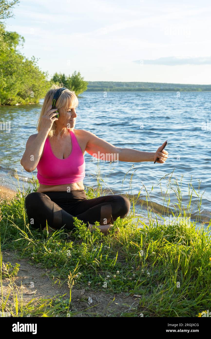 An elderly woman in sports clothes and headphones holds a phone in her hands, watches online exercises for training, waves a greeting with her hand, l Stock Photo