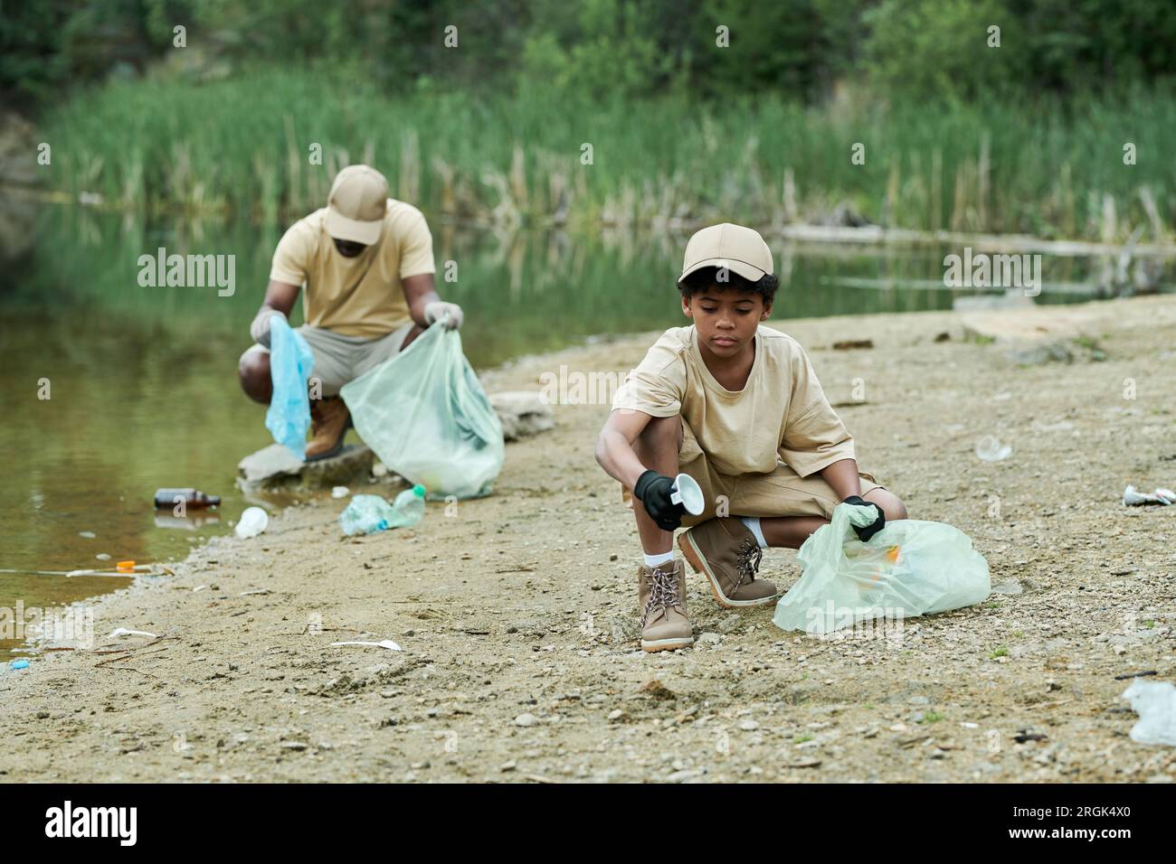 African American child picking up garbage around the lake together with his dad Stock Photo